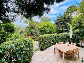 Une terrasse avec une table et des chaises en bois, entourée d’une végétation luxuriante et bordée d’un chemin de pierres.