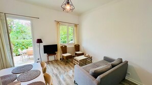 Ein Zimmer mit grauem Sofa, einem Esstisch mit Platzsets, einem Fernseher und einem Fenster mit Vorhängen.