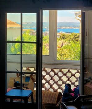 A room with a large window, a wicker chair, a small table, and a blue chair. - The original (Sète)