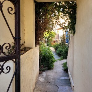 A narrow alleyway with a metal gate, surrounded by greenery and a pathway leading to a building. - The original (Sète)