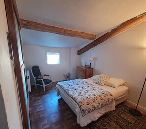 A bedroom with a bed, a chair, a dresser, a lamp, and a window. - The original (Sète)