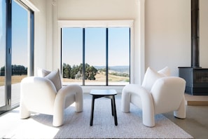 A modern living room with a large window, two white armchairs, a small table, and a black fireplace.