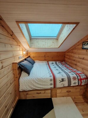 Une petite pièce chaleureuse avec un plafond en pente et un puits de lumière. On y trouve un lit recouvert d’une courtepointe à motifs et agrémenté d’oreillers, des murs en bois et un petit banc en bois.