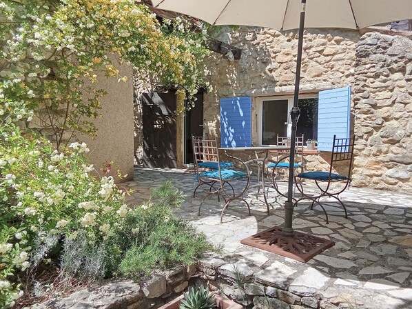 A stone-walled courtyard with a table and chairs, a blue door, and a hanging plant.