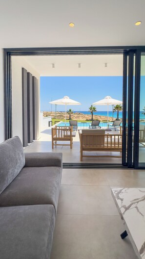 A modern living room with a sofa, a dining table with chairs, and a view of the ocean through a glass door. - Aqua Beachfront Villas #1, Aqua Beachfront Villas - Villa 1 (Ayia Napa)