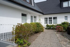 Ein weißes Gebäude mit befestigter Auffahrt, weißem Zaun und verschiedenen Sträuchern und Blumen.