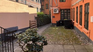 Een smalle steeg met kinderaardewerktegels, een kleine tuin met een metalen bankje, en aangrenzende gebouwen met bakstenen en geverfde gevels.