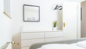 Een slaapkamer met een bed, een kaptafel, een spiegel, een plant en een deur met kapstok.