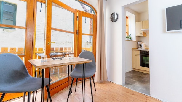 Een kamer met een houten tafel en twee stoelen, een raam met gordijnen, een keuken met kastjes en apparatuur, en een klok aan de muur.
