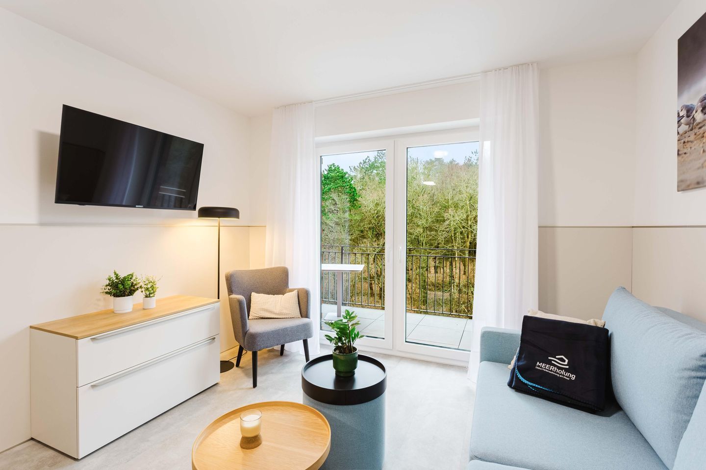 Uma sala de estar moderna com televisão de tela plana, uma poltrona cinza, um sofá, uma mesa de centro e uma varanda com vista para árvores.