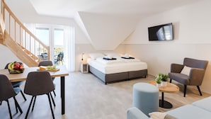A modern, well-lit room with a bed, a dining area with chairs, a small table, a TV mounted on the wall, and a staircase leading to an outdoor area. - MEERholung FeWo 4.10 - Exclusive penthouse with sea view & roof terrace flair - (Cuxhaven)