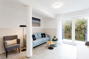 A modern living room with a grey armchair, a light blue sofa, a round coffee table, and a large window with a view of trees. - MEERholung Vacation Rental 1.4 with a balcony and within walking distance of the beach, for 4 people - House (Cuxhaven)