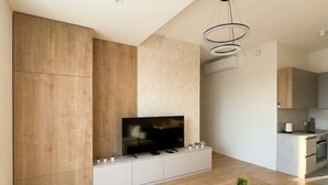 A modern living room with a flat-screen TV, wooden cabinets, a kitchen area, and a dining table. - 8-Min to Airport 1BR & 1LR Flat (Bratislava)