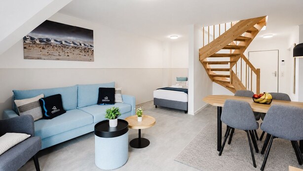 Un espacio de estar moderno con un sofá azul, una mesa redonda, una zona de comedor con mesa y sillas de madera, y una escalera de caracol.