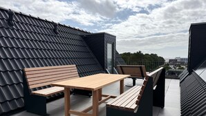 Una terraza en la azotea con una mesa y bancos de madera, vistas de la ciudad y un edificio moderno con tejado inclinado.