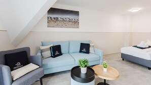 Una habitación de hotel moderna con un sofá azul claro, un sillón gris, una mesita redonda con una planta y una cama con ropa de cama blanca.