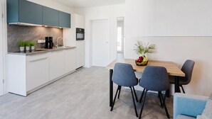 Una cocina moderna con armarios blancos, una campana extractora color verde azulado oscuro y un horno empotrado. Hay una zona de comedor con una mesa de madera y dos sillas grises.