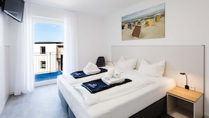 Habitación de hotel con cama, mesita de noche, televisión, un cuadro de una playa y balcón con vista al mar.