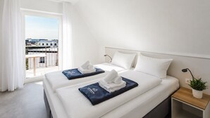 Ein Hotelzimmer mit zwei Betten, einem Balkon, Blick auf Gebäude und einem kleinen Nachttisch mit Pflanze.