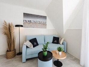 Ein modernes Wohnzimmer mit einem hellblauen Sofa, zwei schwarzen Kissen, einer Zimmerpflanze, einem Holztisch, einer Stehlampe und einem Strandbild an der Wand.