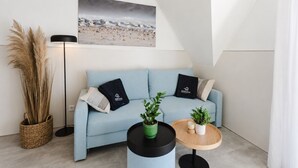 Ein modernes Wohnzimmer mit einem hellblauen Sofa, zwei schwarzen Kissen, einer Zimmerpflanze, einem Holztisch, einer Stehlampe und einem Strandbild an der Wand.