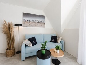 Uma sala de estar moderna com um sofá azul claro, duas almofadas pretas, uma planta verde, uma mesinha de madeira, um abajur e uma pintura com tema de praia na parede.