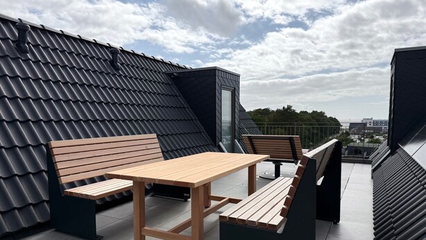 En tagterrasse med et træbord og bænke, udsigt over bybilledet og en moderne bygning med et skråt tag.