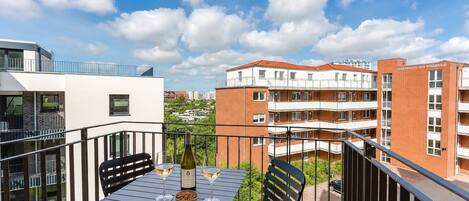 Un balcone con tavolo, due sedie, una bottiglia e bicchieri, affaccia su una zona residenziale con edifici e cielo sereno.