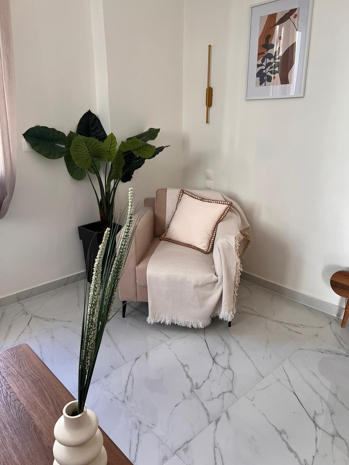 Ein Zimmer mit Marmorboden, einem beige gefärbten Sofa mit weißer Decke und einem Blumentopf sowie einem gerahmten Bild an der Wand.