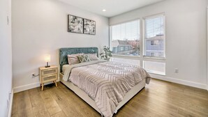 Un dormitorio con cama, mesita de noche, lámpara, obra de arte y ventanas amplias.