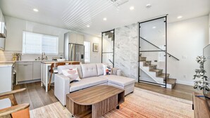 Una sala de estar moderna con un sofá modular, una mesa de centro de madera, una cocina con refrigerador y una escalera con barandas de vidrio.