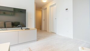 A modern living room with a flat-screen TV, a light-colored wooden floor, white walls, and a hallway leading to a door. - 1BR Apartment Near Airport with Parking (Bratislava)