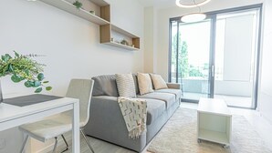A modern living room with a grey sofa, white coffee table, and a desk with a chair. There are shelves with plants and a large window providing a view. - 1BR Apartment Near Airport with Parking (Bratislava)