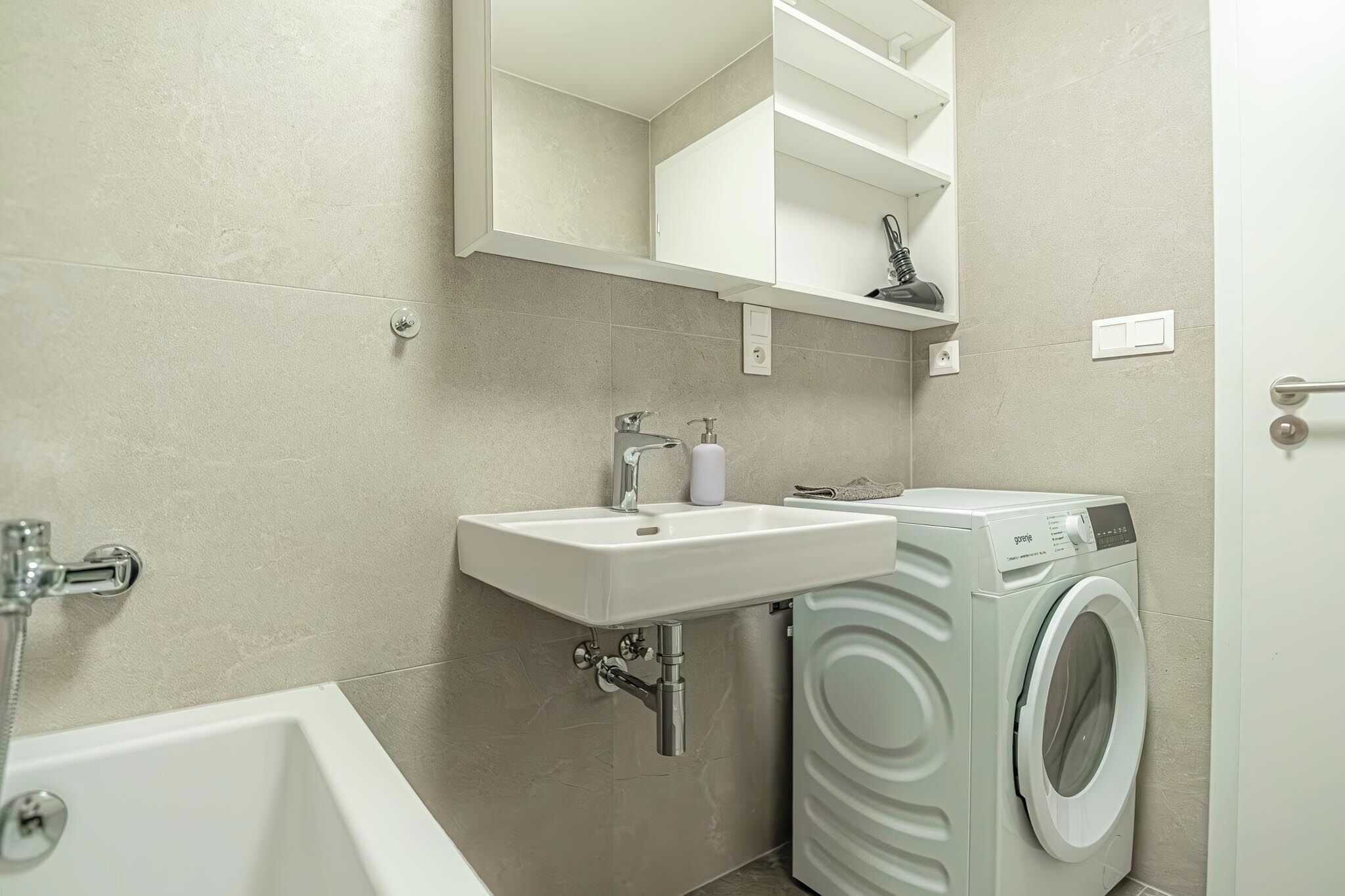 A bathroom with a sink, mirror, shelving, and a washing machine.