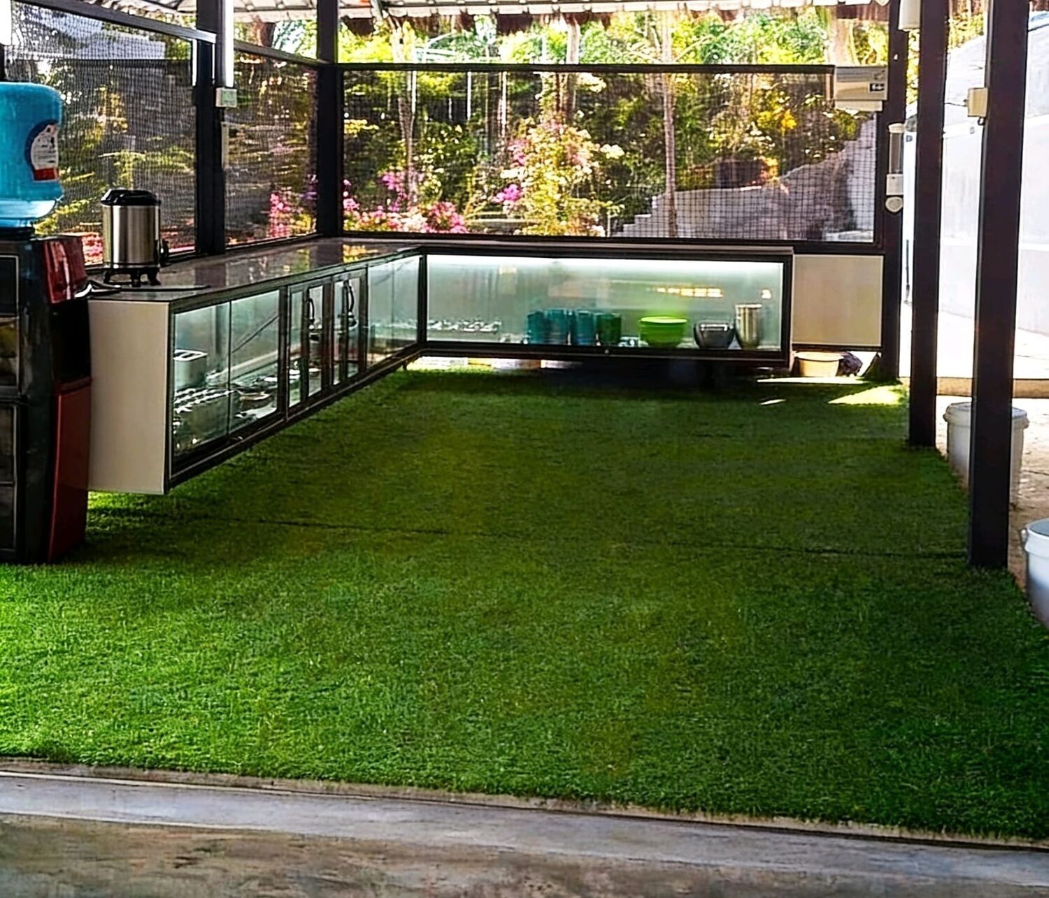 Un patio couvert avec un revêtement de gazon synthétique, une structure en verre et une vue sur un jardin.