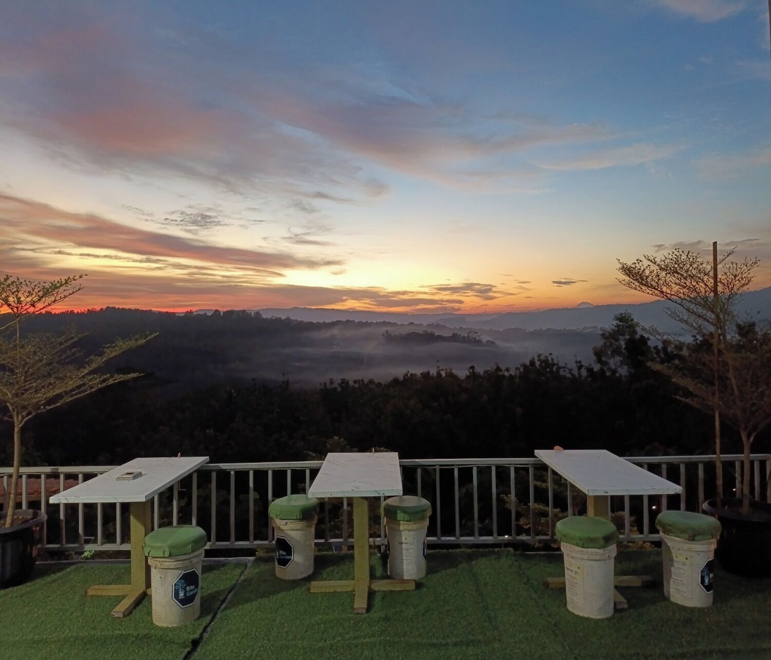 A terrace with a table and chairs, a railing, and a view of a valley during sunset.