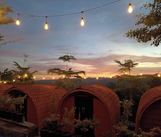 Uma fileira de chalés rústicos com telhados em arco e luzes de corda, com o céu ao pôr do sol ao fundo.