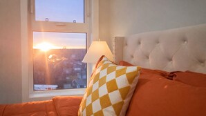 Een bed met een wit gepolijst hoofdbord, een oranje deken en een geel-wit geruit kussen. Een raam toont een uitzicht op een zonsondergang en gebouwen.