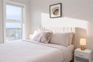 A bedroom with a bed, pillows, a bedside table with a lamp, a framed artwork on the wall, and a window.