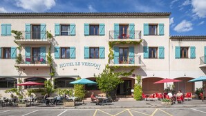 Un hotel con mesas al aire libre, un cartel que dice 'HOTEL LE VERDON' y un cielo azul despejado.
