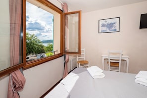 Ein Zimmer mit einem Bett, einem Sessel, einem Tisch, einem Fenster mit Blick auf Grünflächen und ein Gewässer, sowie einem gerahmten Bild an der Wand.