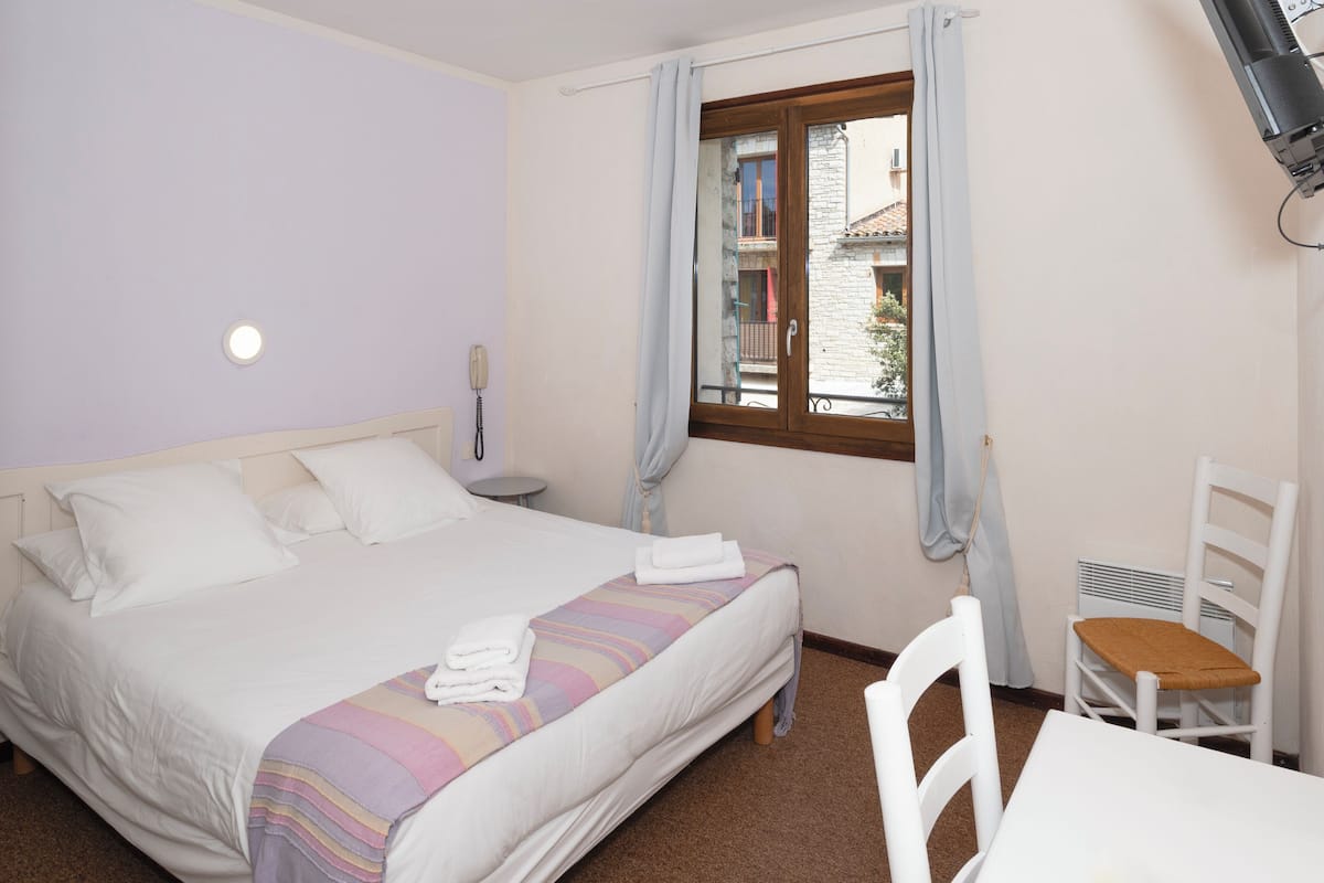 A hotel room with a bed, a bedside table, a chair, a window with curtains, and a view of buildings outside.