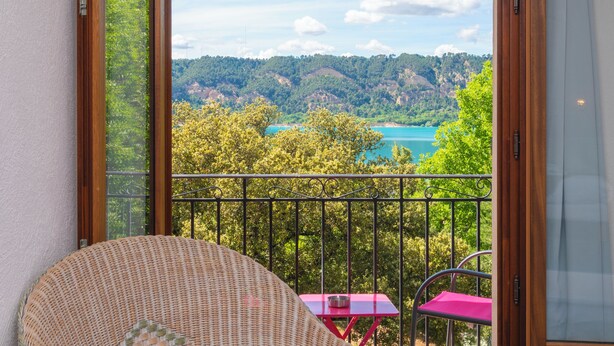 Un balcón con una silla de mimbre, una mesita con mantel rosa y vistas a un lago y un bosque.