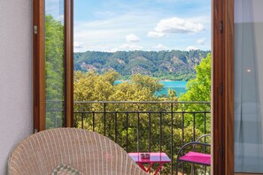 Un balcon avec un fauteuil en osier, une petite table recouverte d’une nappe rose, et une vue sur un lac et une forêt.