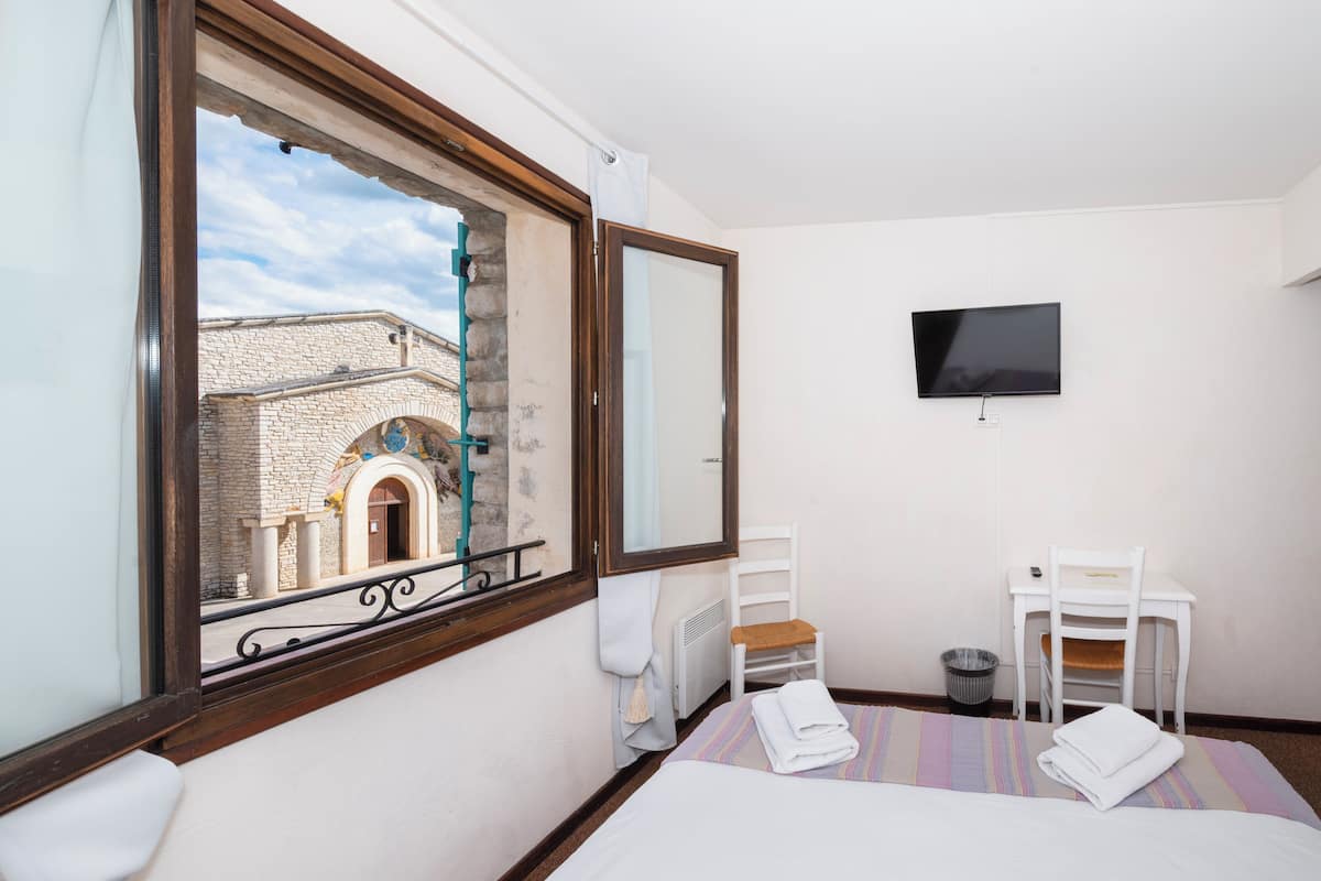 A hotel room with a bed, a desk, a chair, a TV, and a window with a view of a building.