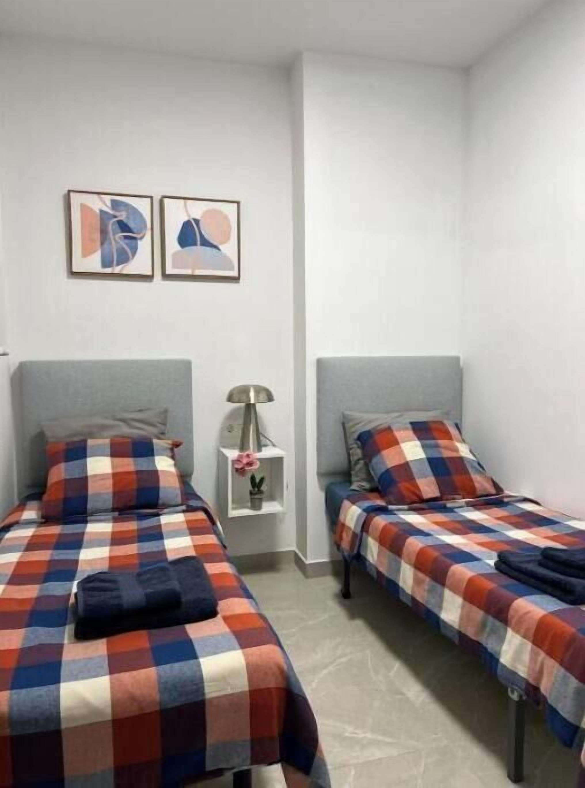 Una habitación con dos camas, cada una con una colcha a cuadros, una mesita de noche con lámpara, y dos cuadros abstractos enmarcados en la pared.