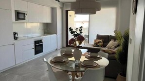 Una cocina moderna con armarios blancos, un microondas y un comedor con mesa de vidrio y sillas blancas.