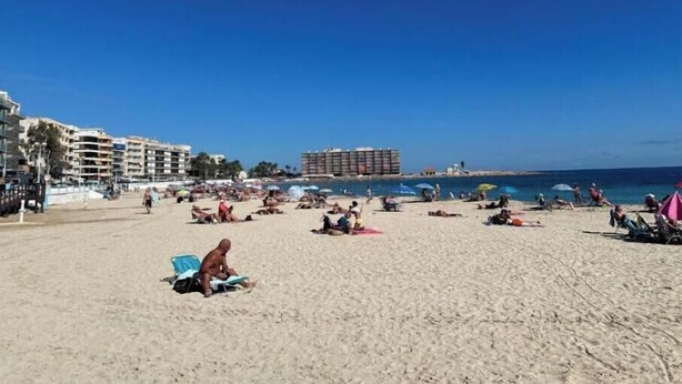 Una playa con gente tomando el sol y nadando, edificios al fondo y un cielo azul despejado.
