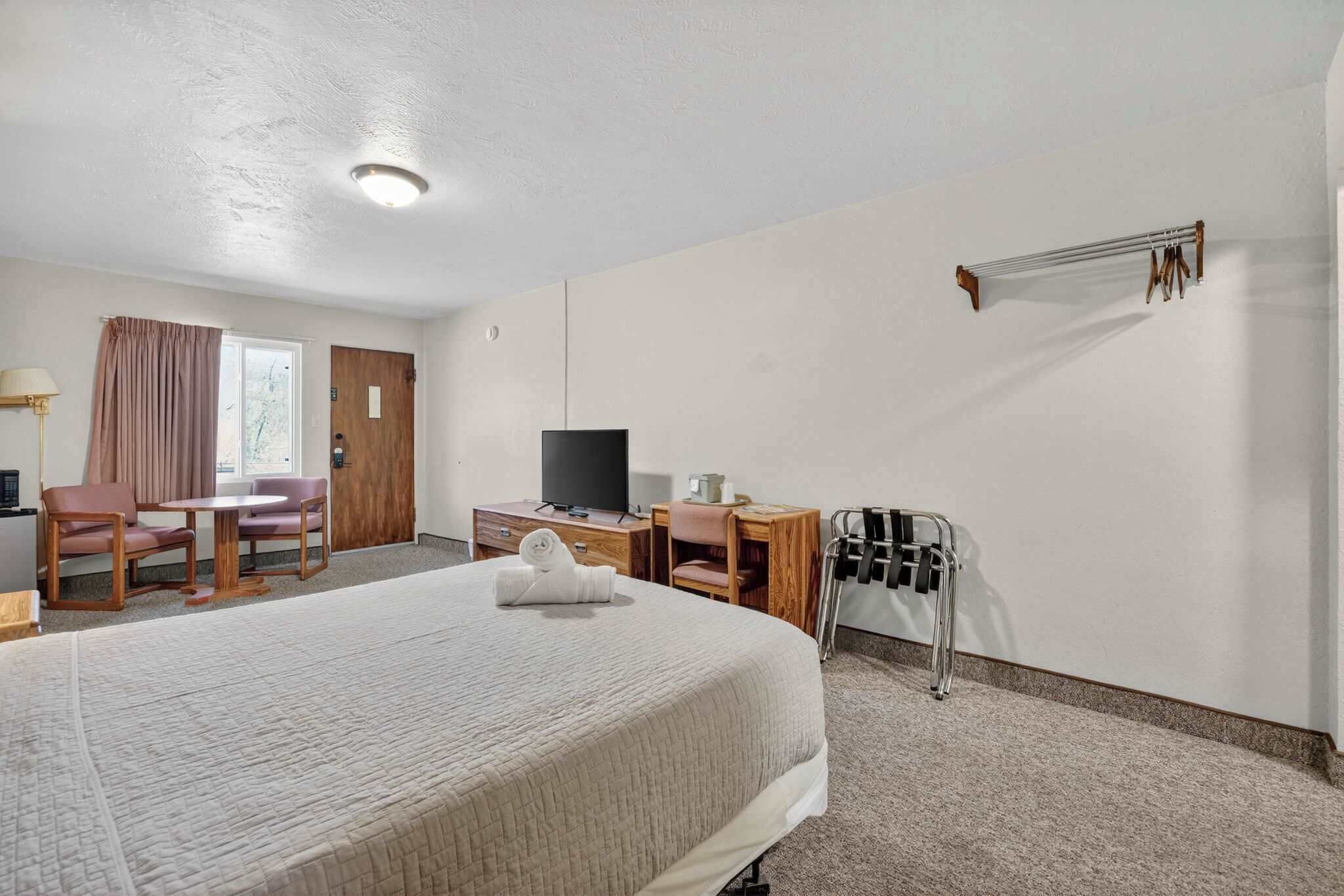 Habitación de hotel con cama, escritorio con computadora, televisión, silla, mesita y ventana con cortinas.