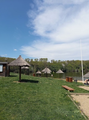 Une zone au bord du lac, avec des parasols en chaume, un chalet en bois et un mât.